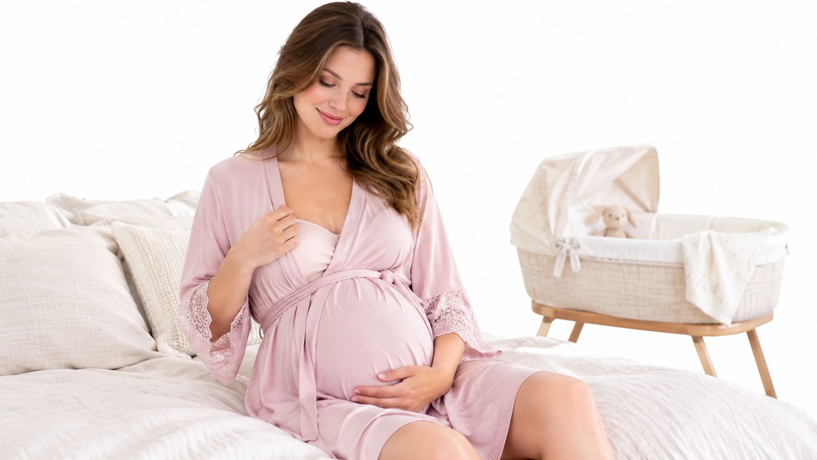 A smiling pregnant woman wearing a soft pink maternity bathrobe sitting on a bed beside a bassinet.