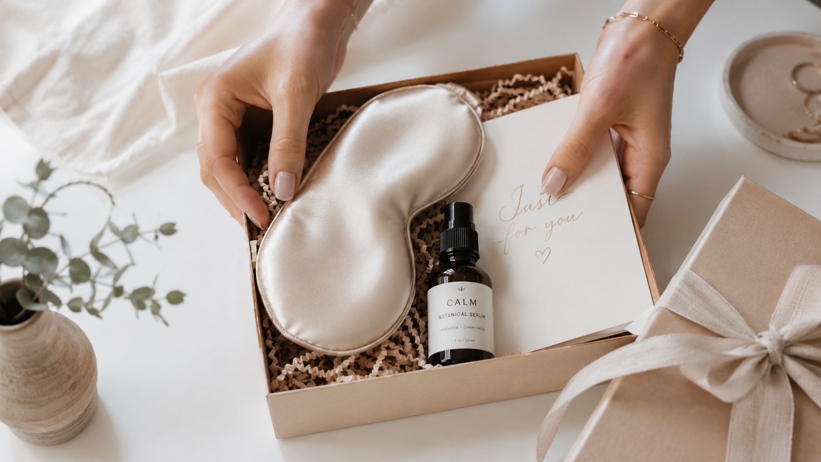 A person holding a gift box containing a silk sleep mask and a bottle of botanical face serum.